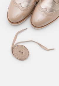 Béžové lakované boty s metalickými zlatými akcenty a vlnitými okraji, doplněné béžovými textilními tkaničkami.