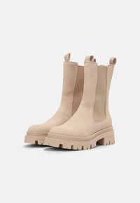 Beige Knöchelstiefel mit elastischen Seitenpaneelen und Zuglaschen, ausgestattet mit einer klobigen Sohle mit geriffelter Textur für verbesserten Halt und Stabilität.