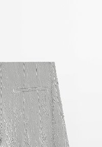 Tissu rayé vertical noir et blanc avec une poche fendue horizontale, posé à plat sur un fond blanc.