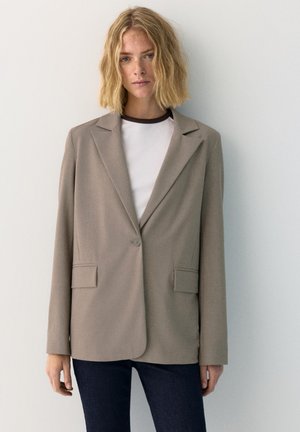 Blonde vrouw met golvend haar, gekleed in een taupekleurig colbert over een wit shirt, staand tegen een effen lichtgrijze muur.