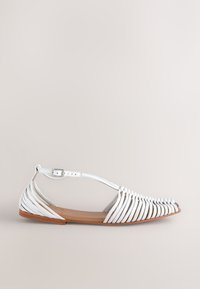 Sandale blanche plate avec un design à brides et une boucle de cheville réglable. Comprend une tige en cuir lisse et une semelle marron flexible.