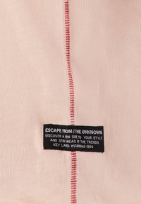 Lichtroze stof met een zichtbare rode gestikte naad. Heeft een zwart label met witte tekst, waarop merkinformatie en een slogan staan.