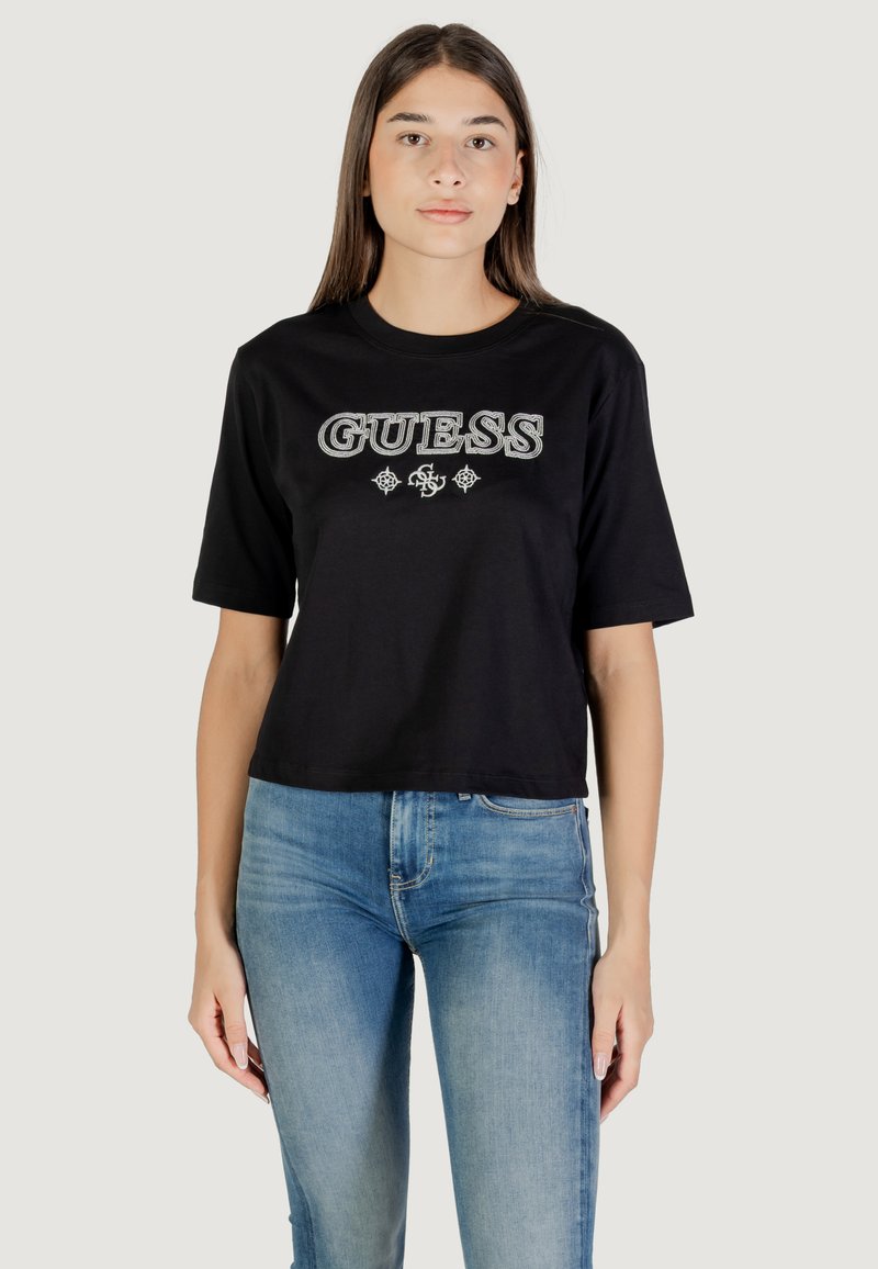 T-shirt corta nera a maniche corte con logo "GUESS" in lettere argentate, indossata con jeans azzurri chiari.