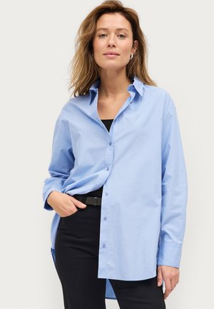 Camisa de botones de manga larga en color azul claro, hecha de algodón, con cuello clásico, bolsillo frontal y corte relajado. Combinada con pantalones negros.