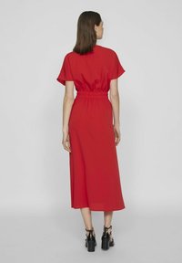 Czerwona midi sukienka z gładkiego materiału, z krótkimi rękawami, elastycznym paskiem w talii i szeroką spódnicą. Widać czarne sandały na słupku.