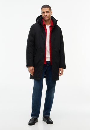 Mann trägt schwarzen Kapuzen-Wintermantel über rotem Hemd, weißem Pullover, blauen Jeans und schwarzen Mokassins, steht vor einfarbig weißem Hintergrund.