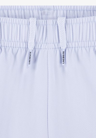 Shorts à taille élastique blanche avec cordons de serrage, présentant un motif en losanges texturé et de petites étiquettes Lacoste sur les deux côtés.