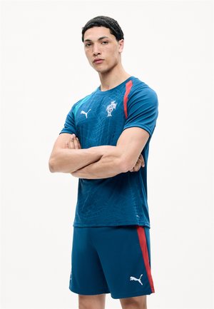 Tânăr stă cu brațele încrucișate purtând echipamentul albastru de fotbal al Portugaliei cu logo-uri Puma și dungă roșie pe pantaloni scurți, pe fundal alb.