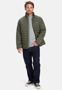 Hombre con cabello castaño claro, vistiendo una chaqueta acolchada de color oliva, suéter gris, jeans oscuros y zapatillas negras, de pie con una mano en el bolsillo.