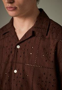 Chemise marron à manches courtes avec broderies florales et découpes. Tissu doux avec fermeture à boutons et col évasé.