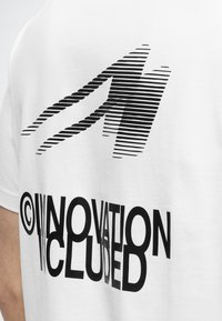 T-shirt blanc avec un graphisme noir du numéro stylisé et incliné "44" au-dessus d’un texte déformé indiquant "INNOVATION EXCLUDED."