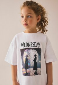 Vit bomull t-shirt med en grafisk tryck som har texten "ONSDAG" och en scen med två figurer framför en spindelnätsdesign.