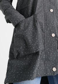 Manteau gris foncé à petits pois blancs, boutons argentés et une main placée dans une poche sur le côté gauche.