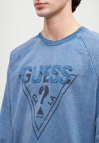 Joven con un sudadera azul que tiene un logo desgastado de Guess con un signo de interrogación dentro de un triángulo.