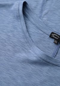 Blauwe katoenen T-shirt met een V-hals, voorzien van een fijne textuur en een subtiel gemêleerd patroon. Het label leest "Superdry Premium," met de beschikbare maten.