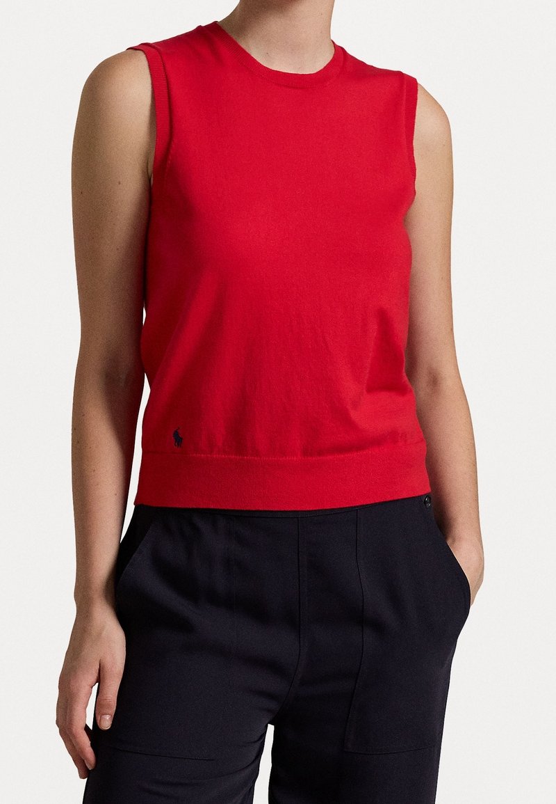 Top sans manches rouge en tissu doux avec un col et un ourlet côtelés. Présente un petit logo près du bas. Porté avec un pantalon foncé.