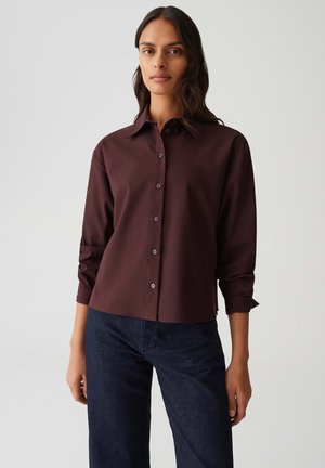 Donna che indossa una camicia button-up bordeaux scuro con maniche lunghe e jeans blu scuri, in piedi contro uno sfondo chiaro e semplice.