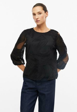 Vrouw met het haar naar achteren gebonden, draagt een zwarte getextureerde doorschijnende blouse met lange mouwen en donkerblauwe spijkerbroek, staand met handen achter haar rug.