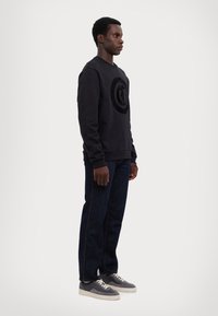 Felpa nera con logo circolare a texture sul davanti, abbinata a jeans di denim scuro e sneakers grigie con dettagli beige.