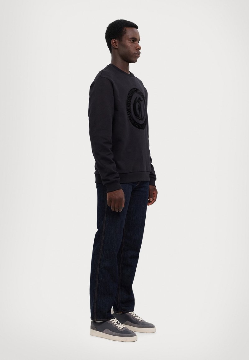 Felpa nera con logo circolare a texture sul davanti, abbinata a jeans di denim scuro e sneakers grigie con dettagli beige.