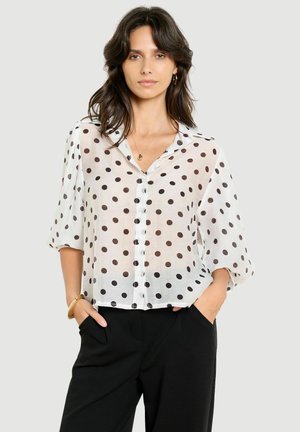 Mujer con cabello oscuro que lleva una blusa blanca transparente con lunares negros y pantalones negros, de pie con las manos en los bolsillos frente a un fondo liso.