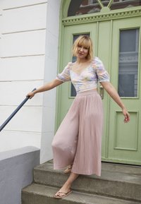 Top leve e floral com mangas bufantes e decote em V, combinado com calças largas plissadas em tom de rosa. Sandálias minimalistas e uma porta verde ao fundo.
