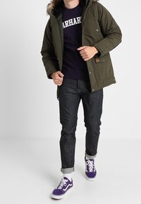 Zelená polstrovaná bunda s umělou kožešinou na kapuci, námořnická mikina, tmavé džíny a fialové tenisky. Disponuje knoflíkovými a zipovými uzávěry, bočními kapsami.