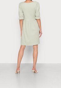 Kobieta stojąca tyłem do kamery, ubrana w jasnozieloną, fakturowaną sukienkę do kolan z bufiastymi rękawami oraz białe sandały na obcasie.