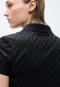 Γυναίκα με κοντά ξανθά μαλλιά που φοράει μαύρο υφασμάτινο πουκάμισο πόλο και μικρά στρογγυλά ασημένια σκουλαρίκια, στραμμένη προς τα αριστερά με φόντο ουδέτερο.