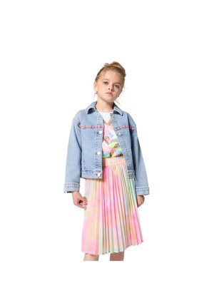 Κορίτσι που φοράει ένα ανοιχτό μπλε τζιν μπουφάν πάνω από μια παστέλ πολύχρωμη πλισέ φούστα και ένα γραφικό μπλουζάκι, στέκεται μπροστά σε ένα λευκό φόντο.