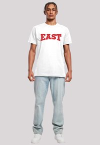 Bílý bavlněný tričko s červenou grafikou "EAST", spárované s světle modrými džínami a bílými teniskami. Jednoduchý design, uvolněný střih.