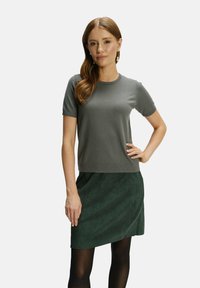 Graues Kurzarmoberteil mit figurbetontem Design und geripptem Halsausschnitt, kombiniert mit einem dunkelgrünen Wildlederrock und schwarzen Strumpfhosen.