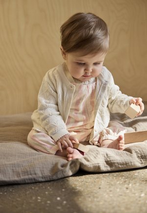 Bambino piccolo seduto su una coperta morbida, indossa un cardigan bianco e un completo a righe rosa, tiene in mano e gioca con blocchi di legno.