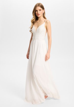 Jeune femme aux longs cheveux blonds portant une robe blanche sans manches avec un corsage texturé et une jupe fluide sur un fond clair uni.