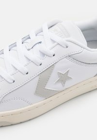 White leather sneaker with textured surface, silver star detail, and beige sole. Features white laces and padded tongue for comfort.