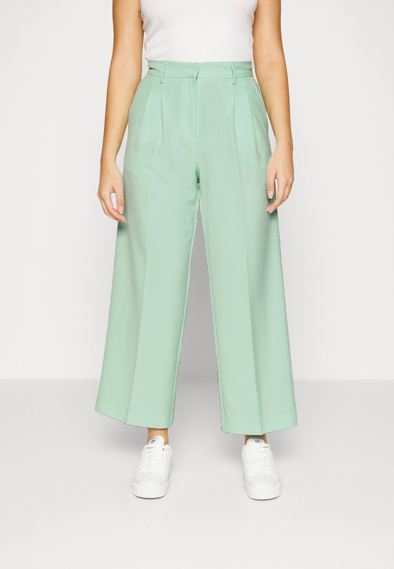 Pantalons à jambes larges mint en tissu lisse, avec des plis à l'avant et une taille ajustée, associés à des baskets blanches.