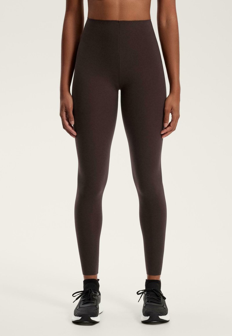 Hochgeschnittene Leggings aus dunkelbraunem Stoff, eng anliegend mit glatter Textur, ausgestattet mit einem gesteppten Bund und kombiniert mit schwarzen Sportschuhen.