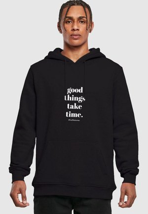Schwarzer Hoodie aus Baumwolle mit einer Kängurutasche. Auf der Vorderseite steht in weißem Text "Gute Dinge brauchen Zeit. #VertraueDerReise."