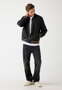 Jachetă bomber din material matlasat, de culoare neagră, cu manșete și tiv cu dungi aurii, fermoar în față și buzunare laterale. Purtați-o peste un tricou alb și asortați-o cu blugi negri.