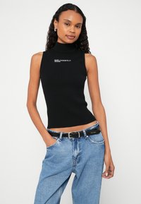 Top preto sem mangas com acabamento em canelado e gola alta, com logótipo branco "KARL LAGERFELD JEANS." Combinado com jeans azul claro de cintura alta e cinto preto.