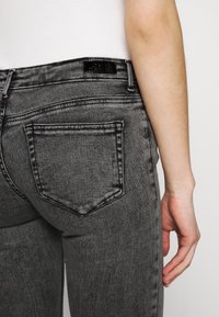 Jeans ajustados de color gris oscuro con una cintura alta, que cuentan con bolsillos traseros y una pequeña etiqueta negra y brillante en la cintura. Textura de denim suave.