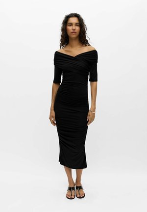 Frau mit lockigem Haar, die ein tailliertes schwarzes off-shoulder Midi-Kleid und schwarze Sandalen trägt, steht vor einem schlichten weißen Hintergrund.