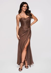 Femme souriante portant une robe marron à bretelles froncées avec une fente haute sur la cuisse et des talons assortis à bride à la cheville, posant la main sur la hanche.
