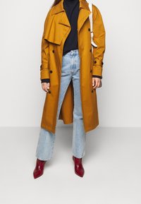 Musztardowy trencz z czarnymi guzikami, zestawiony z jasnoniebieskimi dżinsami i błyszczącymi czerwonymi botkami na szpic; pod spodem czarny golf.