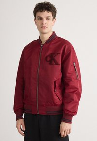 Rode bomberjack met een rits aan de voorkant, geribbelde kraag en manchetten. Heeft geborduurd "CK"-logo en zijzakken. Glad, glanzend stoftextuur.
