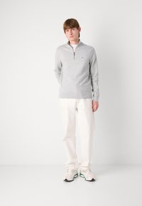 Pulover gri deschis cu un design cu fermoar pe jumătate, textură tricotată și un logo pe piept, asortat cu pantalonii albi și teniși cu un model.