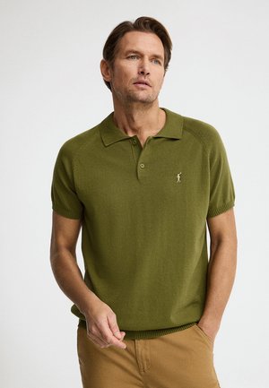 Mężczyzna o jasnobrązowych włosach, ubrany w oliwkowozieloną, krótką, dzianinową koszulkę polo i camelowe spodnie, stojący na jednolitym tle.