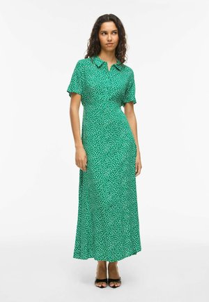 Frau trägt ein grünes, kurzärmeliges Maxikleid mit Kragen und weißen Punkten, steht mit locker an den Seiten hängenden Händen vor weißem Hintergrund.