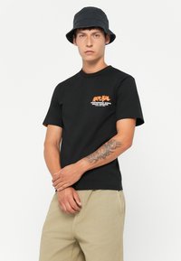Young man wearing a black bucket hat, black graphic t-shirt with orange text, beige pants, and a floral tattoo on his left forearm.