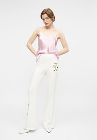 Γυναίκα όρθια με τα χέρια στις τσέπες, φοράει ροζ σατέν καμιζόλα και άσπρο καμπάνα παντελόνι με κεντήματα λουλουδιών.